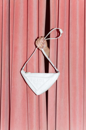 Mão elegante surge entre cortinas de veludo rosa, exibindo bolsa triangular branca da Prada — destaque entre as melhores marcas de bolsas femininas pela sofisticação, design moderno e luxo evidente.