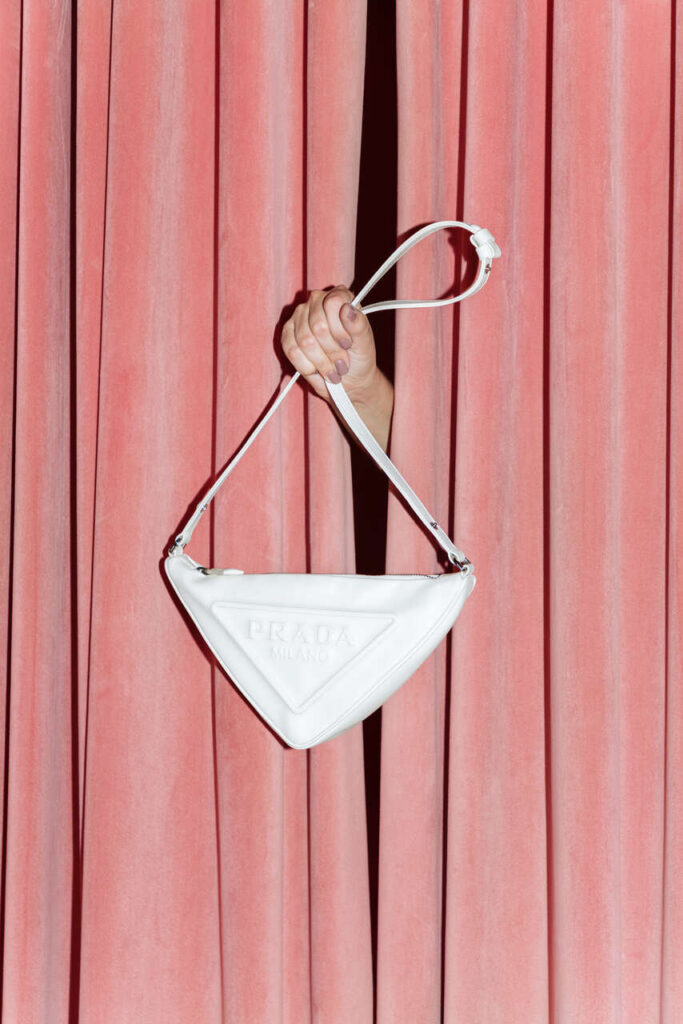 Mão elegante surge entre cortinas de veludo rosa, exibindo bolsa triangular branca da Prada — destaque entre as melhores marcas de bolsas femininas pela sofisticação, design moderno e luxo evidente.