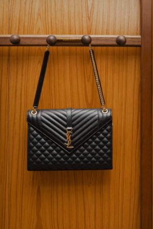 Bolsa preta YSL com logo dourado e alça de corrente pendurada em suporte de madeira — ícone das melhores marcas de bolsas femininas, unindo elegância clássica e luxo contemporâneo.