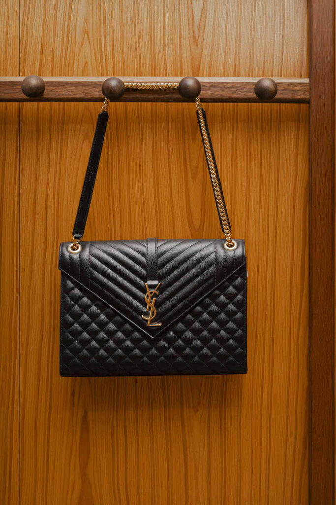 Bolsa preta YSL com logo dourado e alça de corrente pendurada em suporte de madeira — ícone das melhores marcas de bolsas femininas, unindo elegância clássica e luxo contemporâneo.