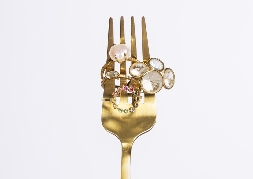 Um garfo dourado sustenta vários anéis decorativos com pérolas e pedras preciosas, lembrando coleções exclusivas de marcas de joias de luxo brasileiras em composição artística.