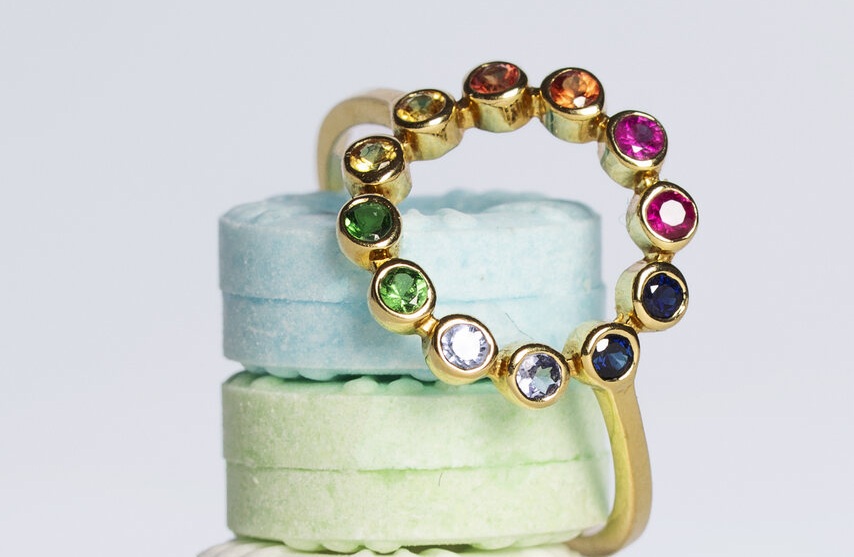 Um anel dourado com gemas coloridas repousa sobre doces pastel, criando contraste vibrante inspirado em campanhas criativas de marcas de joias de luxo brasileiras renomadas.