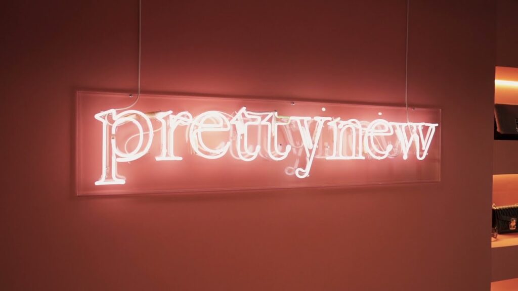Letreiro neon ‘Prettynew’ em loja de revenda de moda sustentável, destacando tendência de reutilização e second hand no varejo fashion.