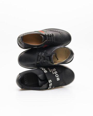Três sapatos masculinos de luxo empilhados: brogue clássico com faixa verde-vermelha, social minimalista e tênis moderno com elástico “BOSS” em destaque.